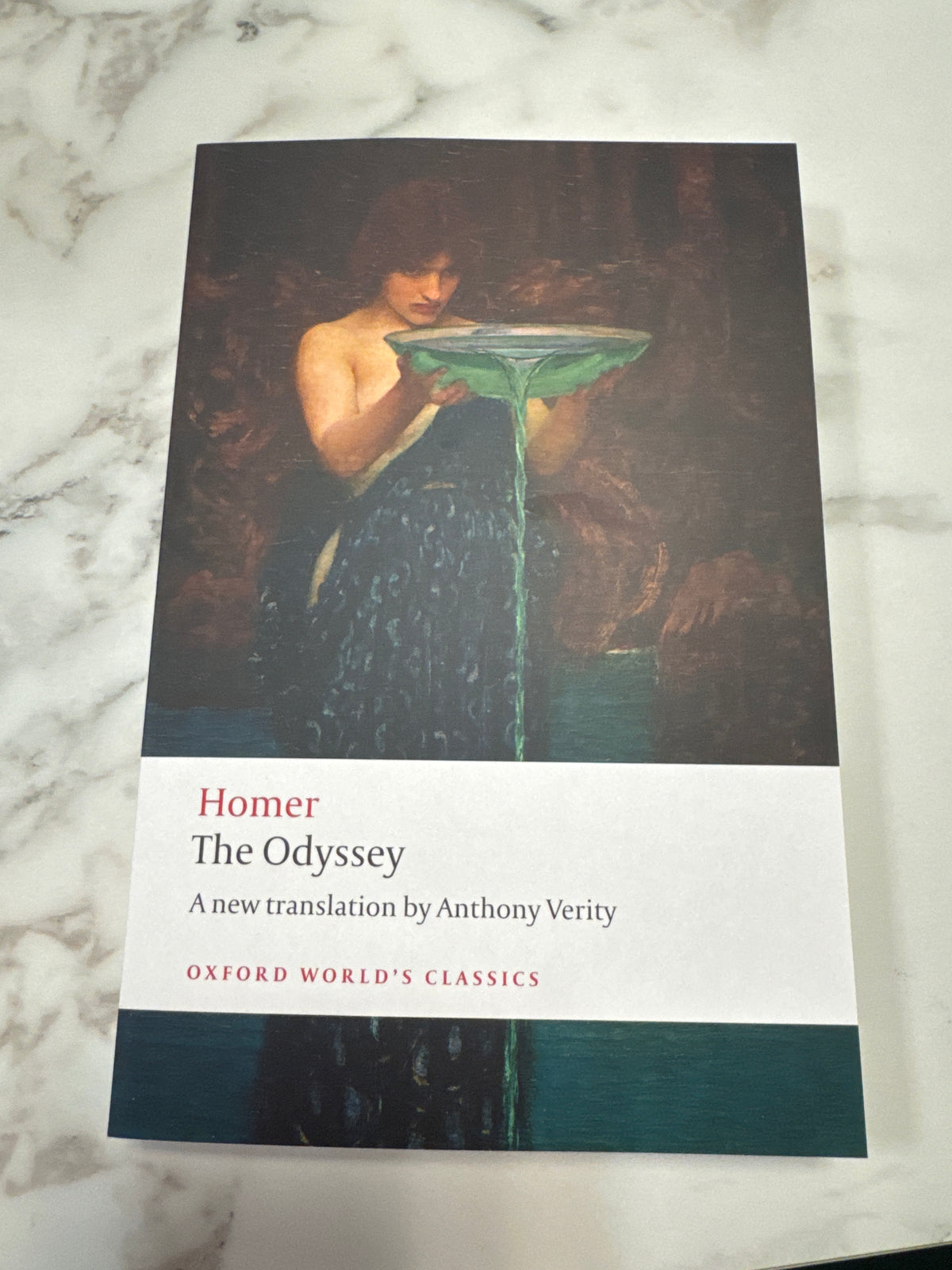 A paperback copy of Homer's The Odyssey from Oxford World's Classics, featuring a dark classical painting on the cover, placed on a white and gray marble surface.