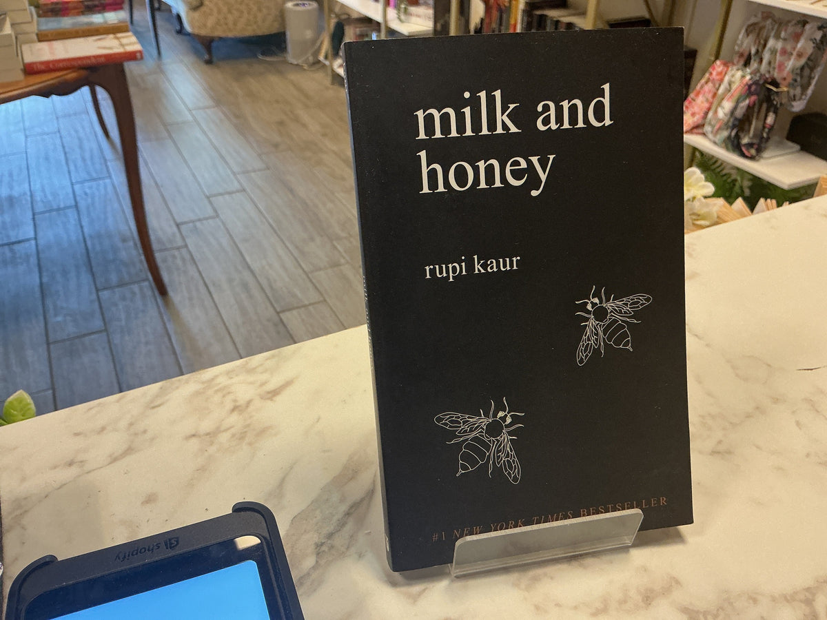 A hardcover book titled 'milk and honey' by Rupi Kaur, standing upright on a clear display stand on a marble counter inside a bookstore.