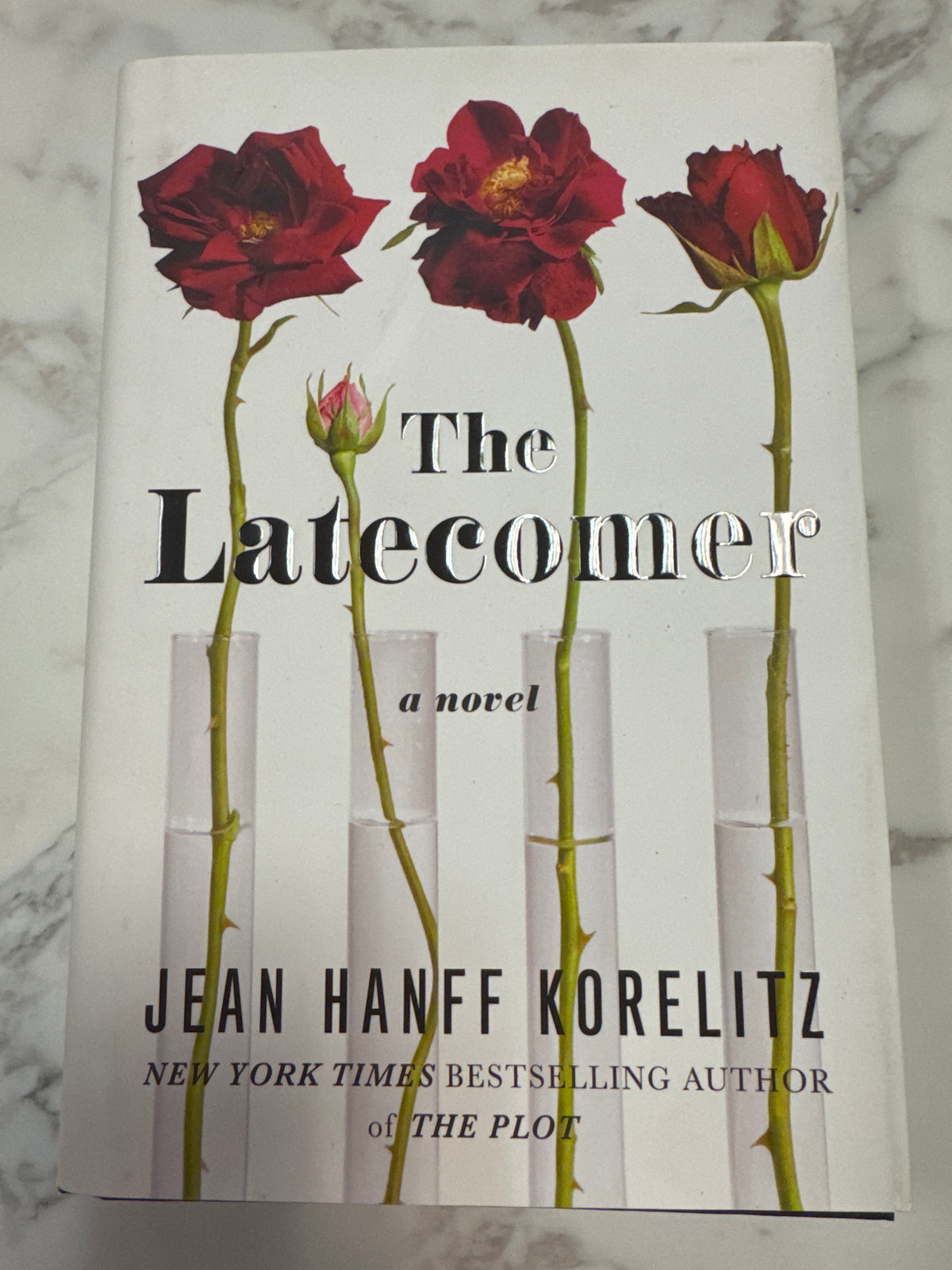 The Latecomer hardcover novel by Jean Hanff Korelitz with red roses in test tubes on cover