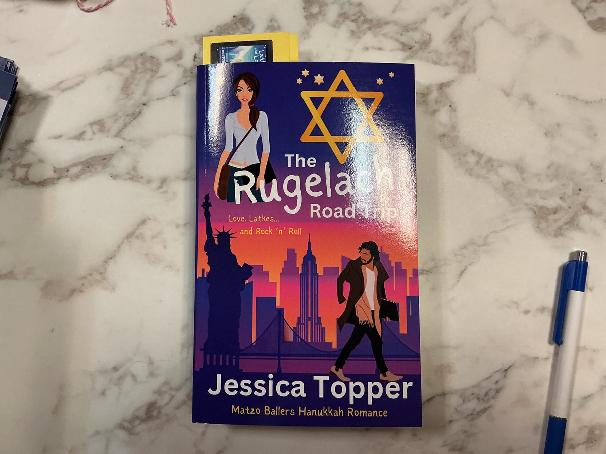 The Rugelach Road Trip paperback book by Jessica Topper with yellow bookmark on marble