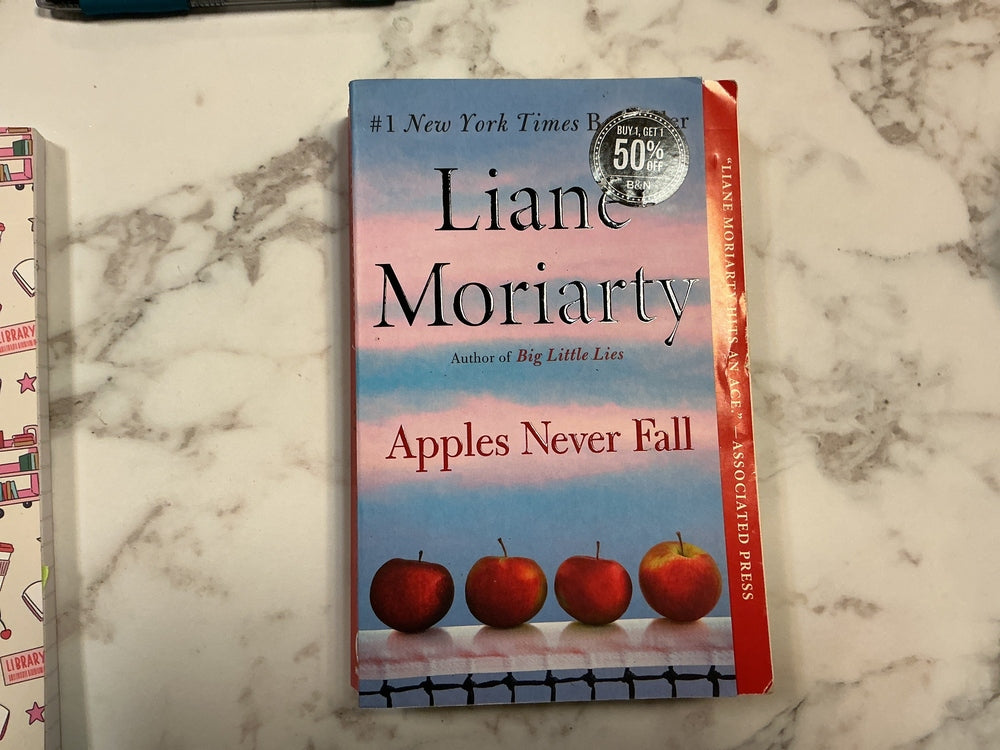 Book titled 'Apples Never Fall' by Liane Moriarty on a marble surface
