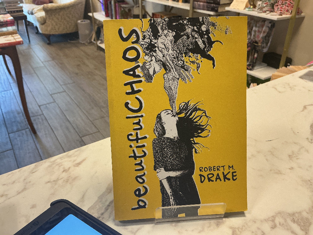 A hardcover book titled 'beautiful CHAOS' by Robert M. Drake, featuring a bright yellow cover with black and white graphic artwork, displayed on a clear acrylic stand on a marble countertop in a store interior.