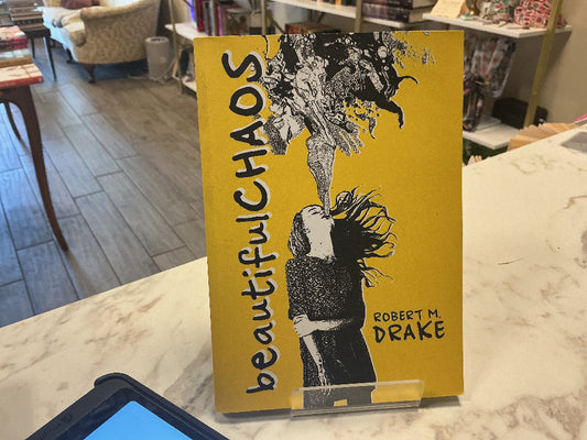A hardcover book titled 'beautiful CHAOS' by Robert M. Drake, featuring a bright yellow cover with black and white graphic artwork, displayed on a clear acrylic stand on a marble countertop in a store interior.