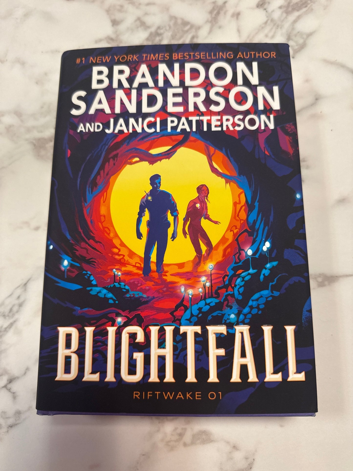 The book cover of 'Blightfall: Riftwake 01' by Brandon Sanderson and Janci Patterson, featuring a dark fantasy illustration of two figures in front of a glowing orange and yellow circular portal, placed on a light marble surface.