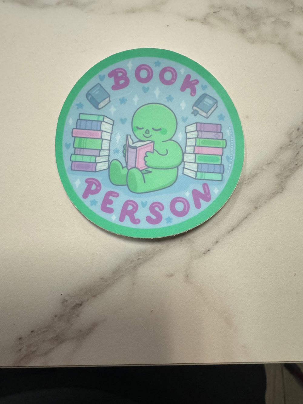 Circular sticker with a green character reading a book, surrounded by text on a marble background