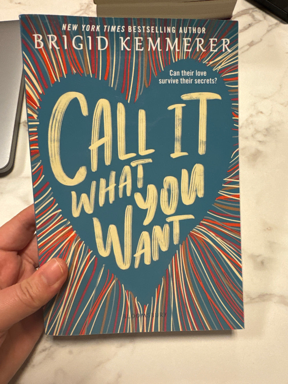 Book titled 'Call It What You Want' by Brigid Kemmerer held by a hand on a marble surface.