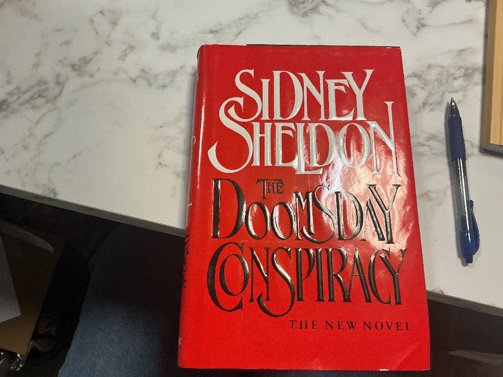 Book titled 'The Doomsday Conspiracy' by Sidney Sheldon on a marble surface with a pen next to it.