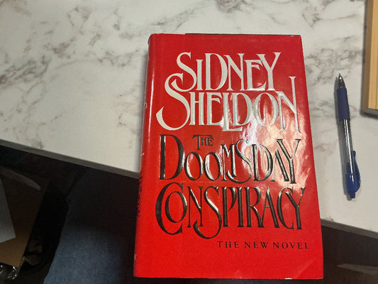 Book titled 'The Doomsday Conspiracy' by Sidney Sheldon on a marble surface with a pen next to it.