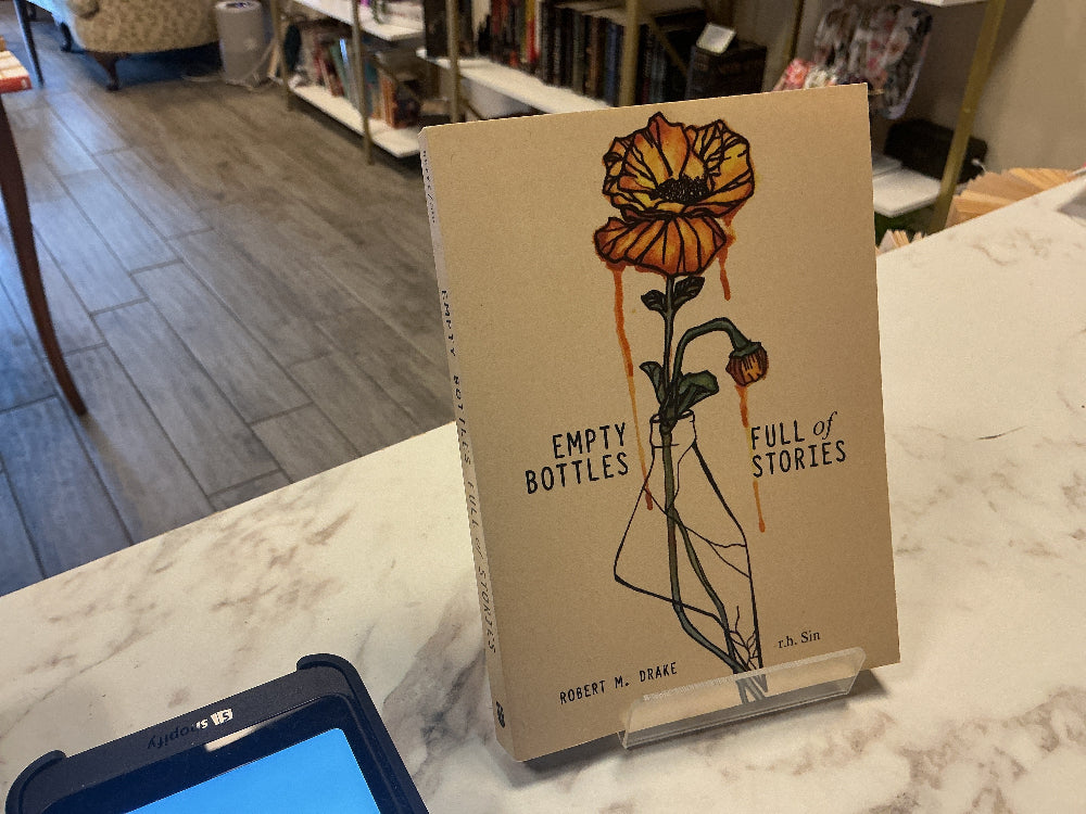 A book titled 'Empty Bottles Full of Stories' by Robert M. Drake, featuring a beige cover with an illustration of a dripping orange flower in a glass bottle, displayed on a marble counter in a bookstore.