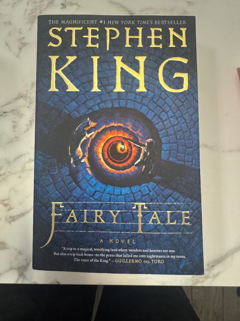 Stephen King's 'Fairy Tale' novel book cover, featuring a dark blue background with a glowing orange spiral and gold lettering, photographed on a white marble surface.
