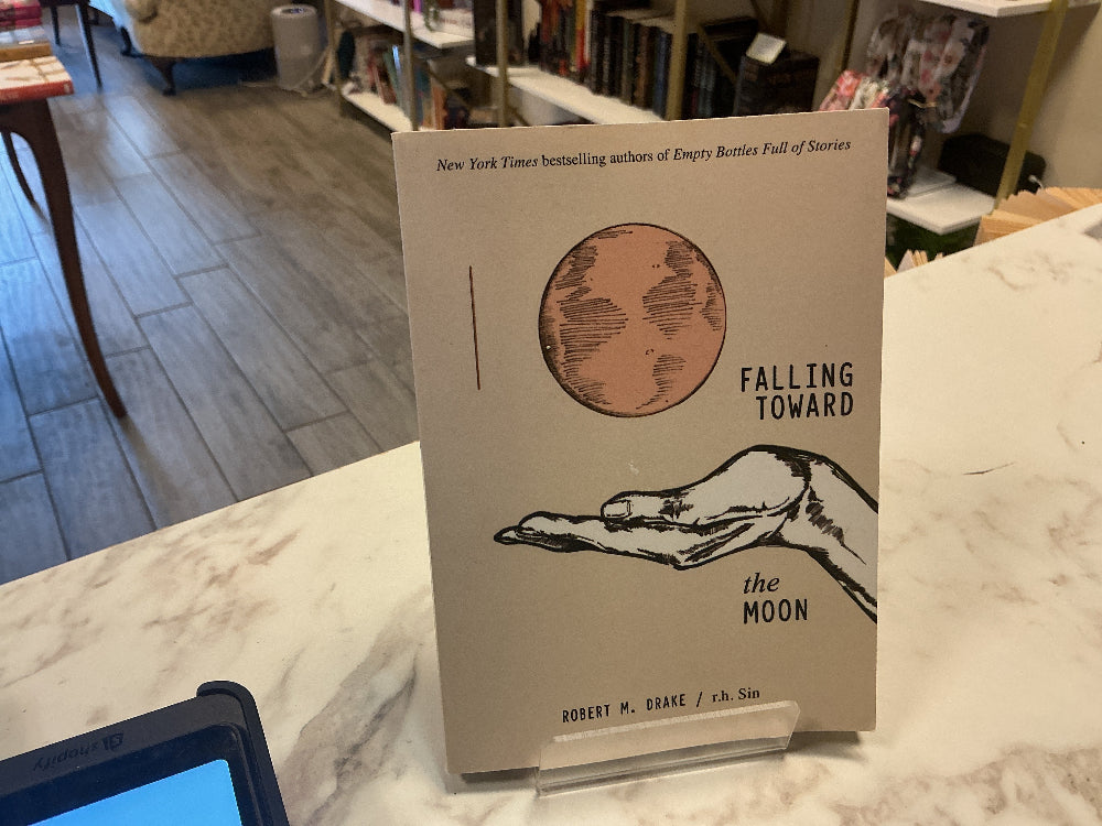 A book titled 'Falling Toward the Moon' by Robert M. Drake stands upright on a clear display stand on a marble counter inside a bookstore.
