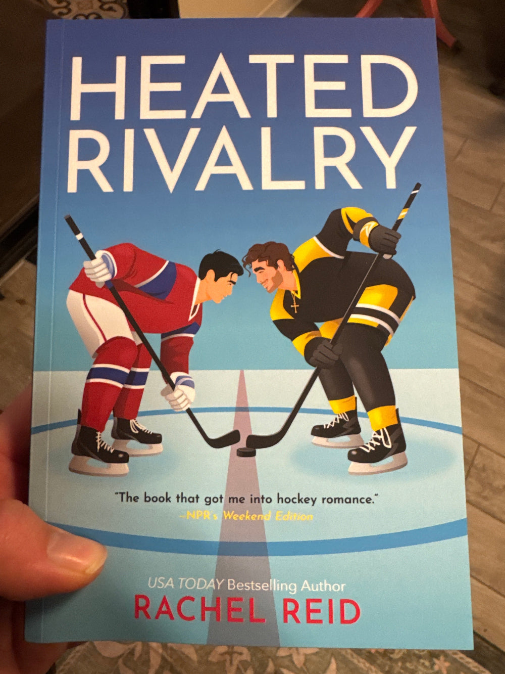 Book cover of 'Heated Rivalry' by Rachel Reid held in a hand.