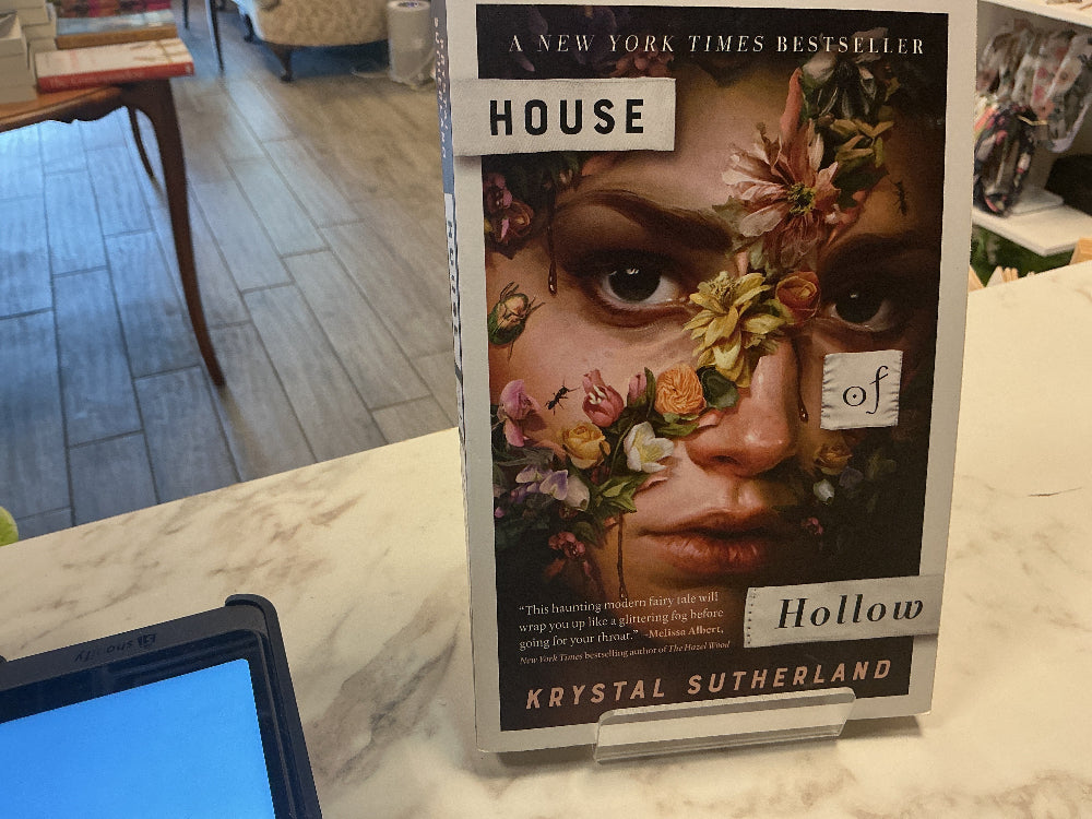 A copy of the book 'House of Hollow' by Krystal Sutherland displayed on a marble counter in a bookstore, with a tablet in the foreground and the store interior blurred in the background.