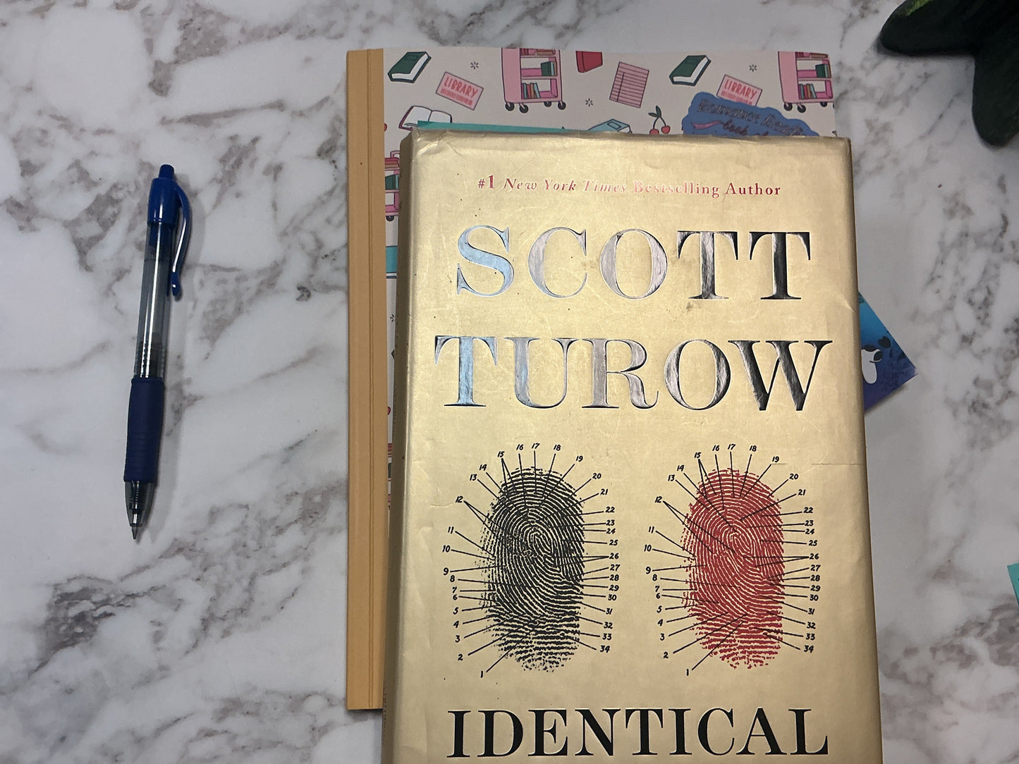 Book titled 'Identical' by Scott Turow on a marble surface with a pen and other items.