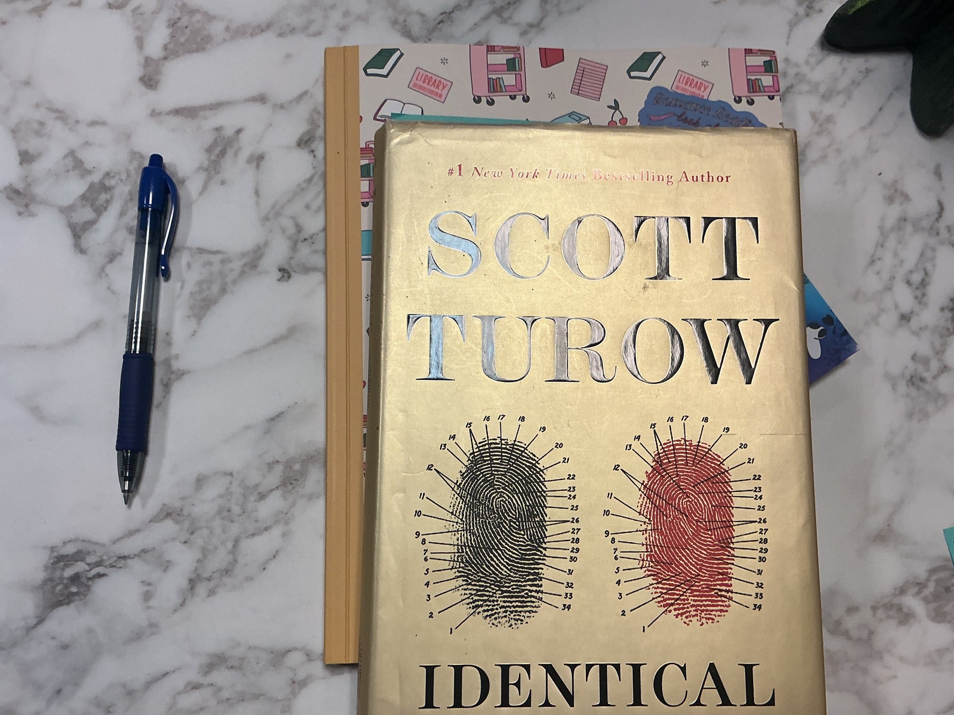 Book titled 'Identical' by Scott Turow on a marble surface with a pen and other items.
