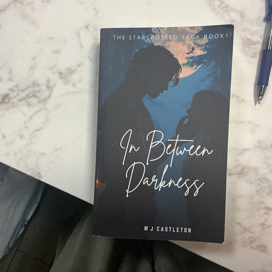 Book titled 'In Between Darkness' by M.J. Castleton on a marble surface with a pen.