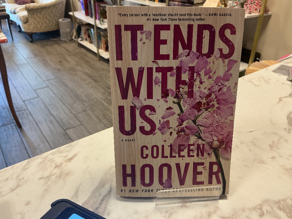 The book 'It Ends With Us' by Colleen Hoover, standing upright on a marble counter in a bookstore. The cover features a light beige background with large dark purple text and pink cherry blossoms.