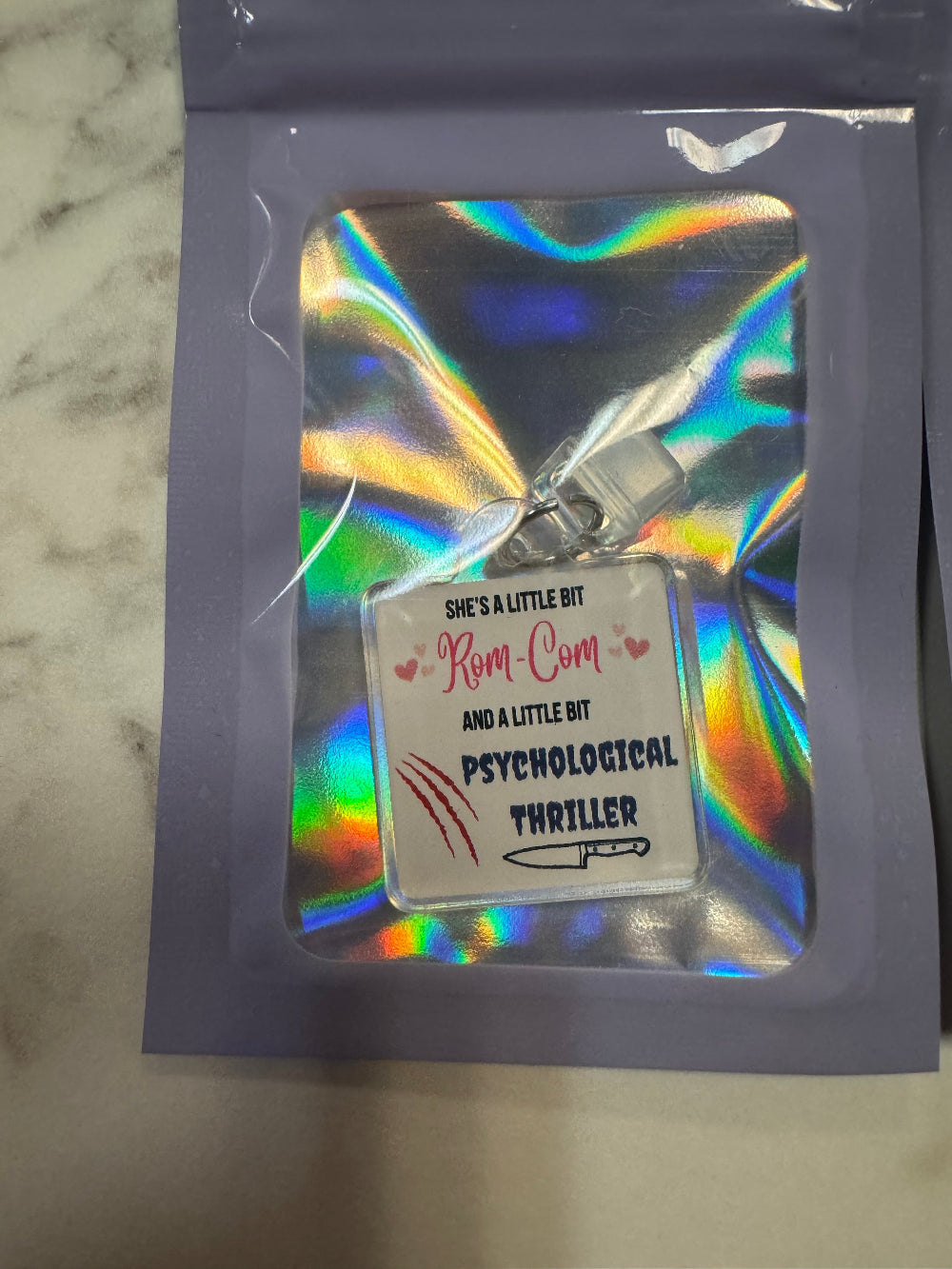 Holographic packaging with kindle charm that says she's a little bit rom-com and a little bit psychological thriller