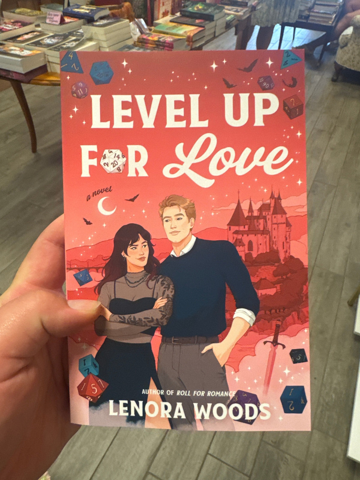 A hand holds a paperback copy of the novel 'Level Up for Love' by Lenora Woods. The book cover features a vibrant red and pink background with an illustration of a young couple, a castle, and floating gaming dice.