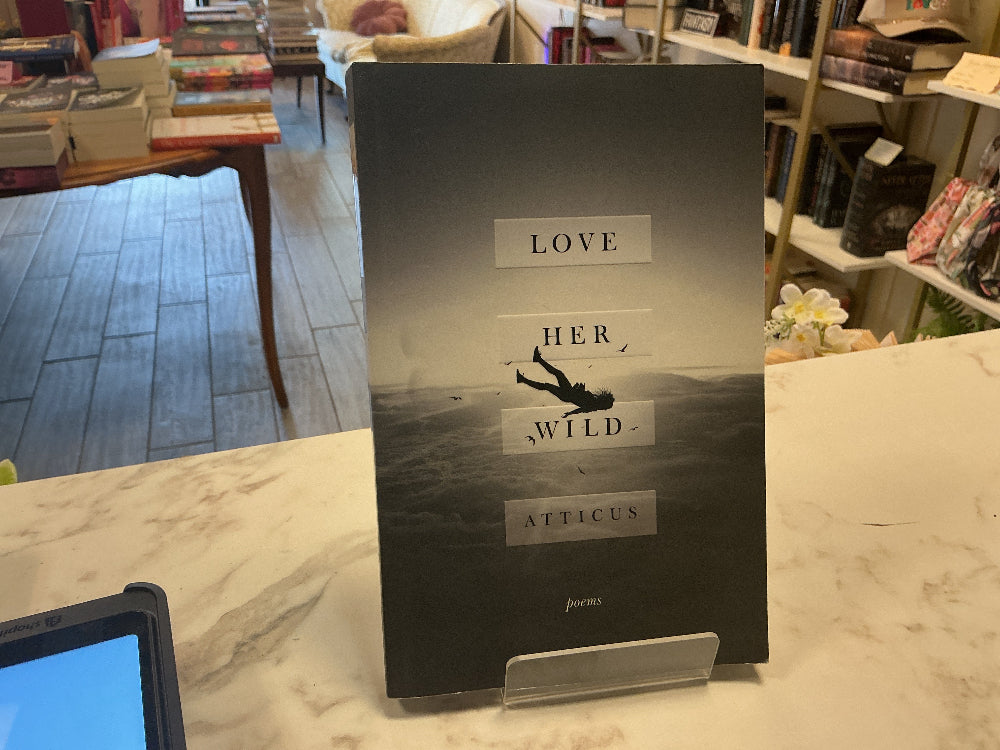 A hardcover book titled 'Love Her Wild' by Atticus stands upright on a clear display stand on a marble counter inside a bookstore. The cover is black and white with a silhouette of a person flying or falling, and the title is displayed in white rectangular blocks.