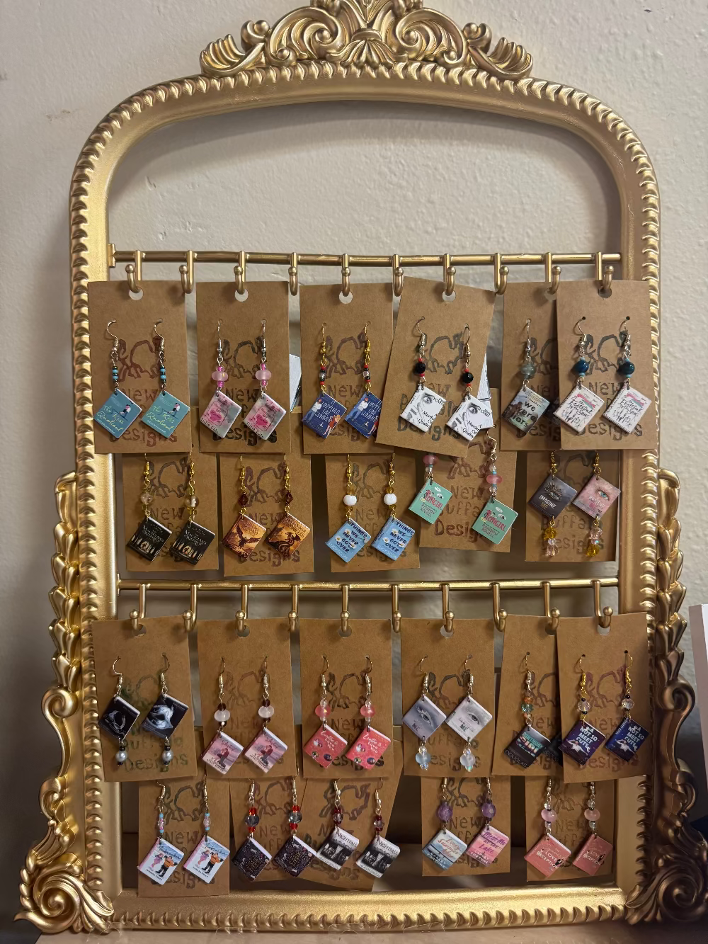 A gold ornate mirror frame used as a jewelry display stand, holding multiple pairs of handmade earrings on brown paper tags against a light beige wall.