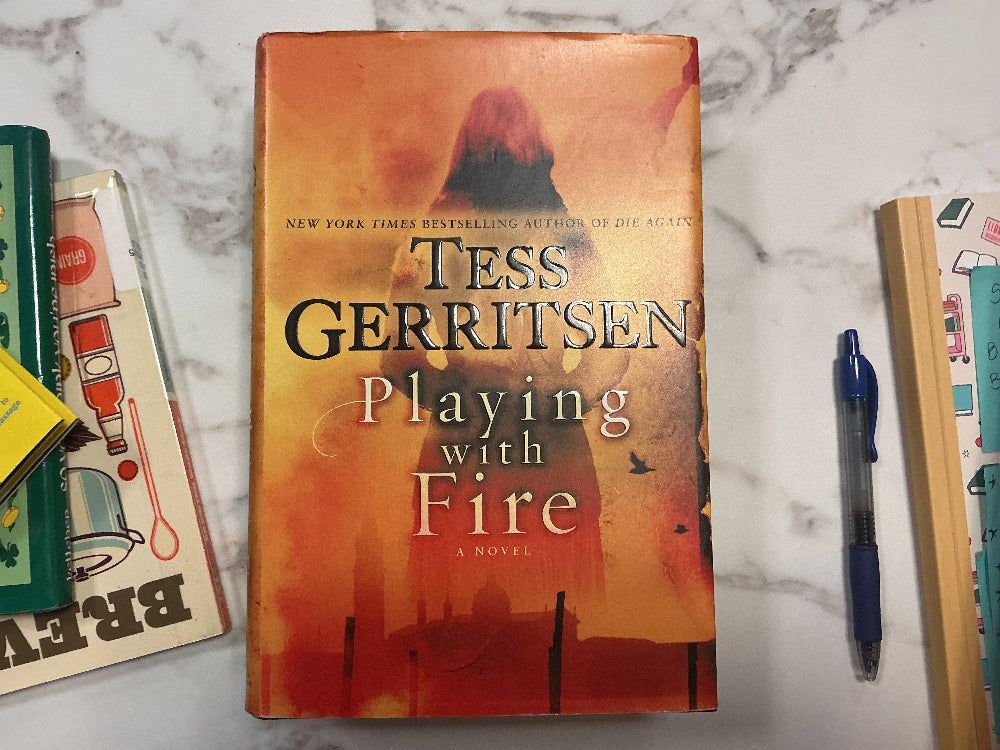 Book titled 'Playing with Fire' by Tess Gerritsen on a marble surface with other books and a pen.