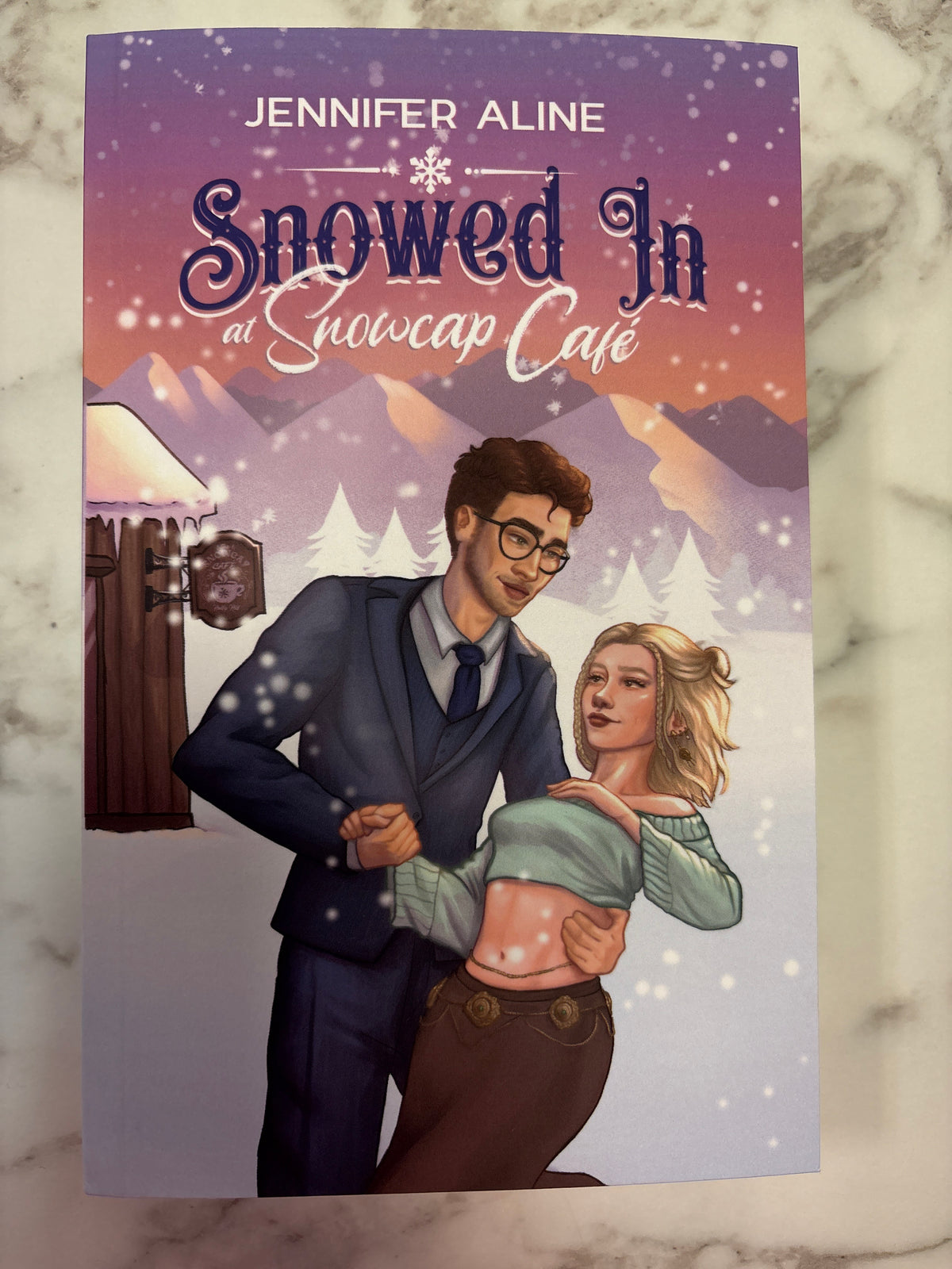 Snowed In at Snowcap Cafe romance novel by Jennifer Aline with illustrated couple in snowy landscape