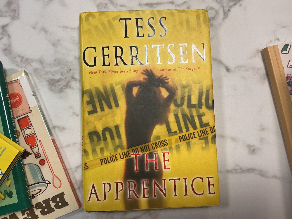 Book titled 'The Apprentice' by Tess Gerritsen on a marble surface with other books.