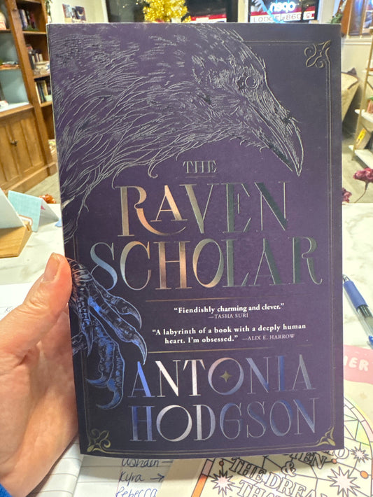 Book titled 'The Raven Scholar' by Antonia Hodgson held in a hand with a blurred background.
