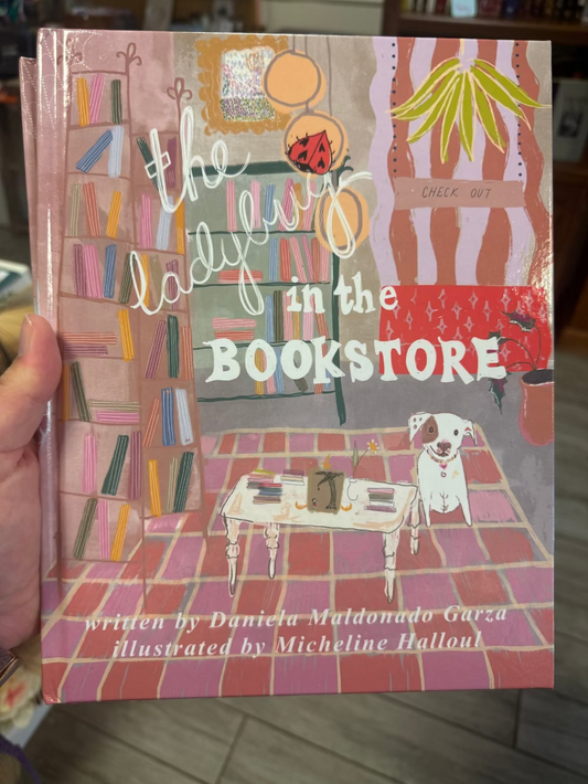 A hand holds a hardcover book titled "The Ladies in the Bookstore" with colorful illustrated cover art showing bookshelves, a dog, and a table. The cover has pink, orange, green, and white tones, and the text at the bottom credits Daniela Maldonado Garza as the writer and Micheline Halloul as the illustrator.