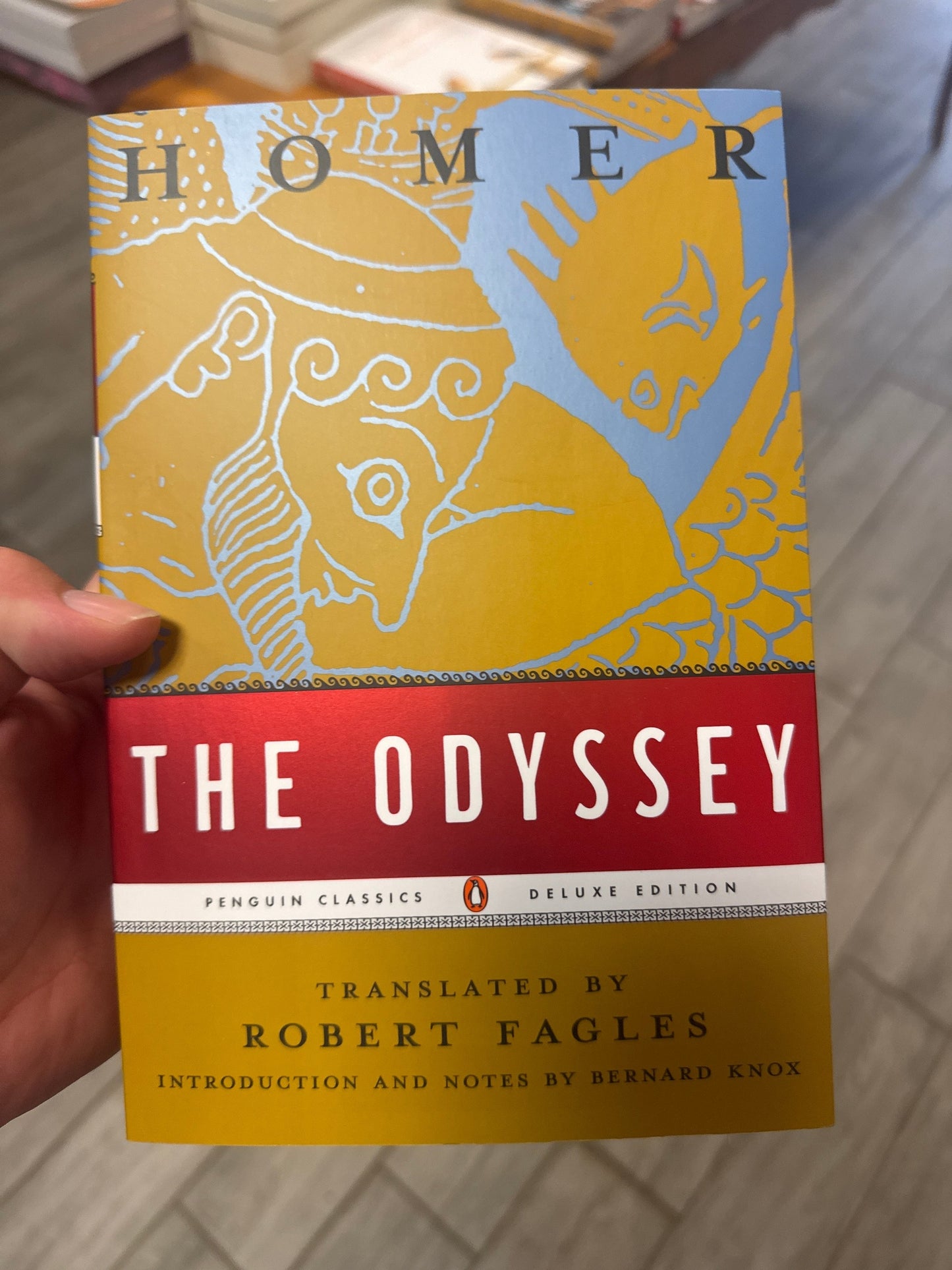 A hand holding a Penguin Classics Deluxe Edition paperback of Homer's The Odyssey, showing the front cover with its yellow, red, and blue illustration against a blurred indoor background.
