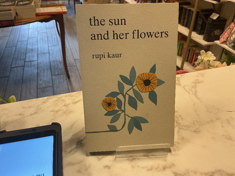 The book 'The Sun and Her Flowers' by Rupi Kaur displayed on a clear acrylic stand on a marble counter inside a bookstore.