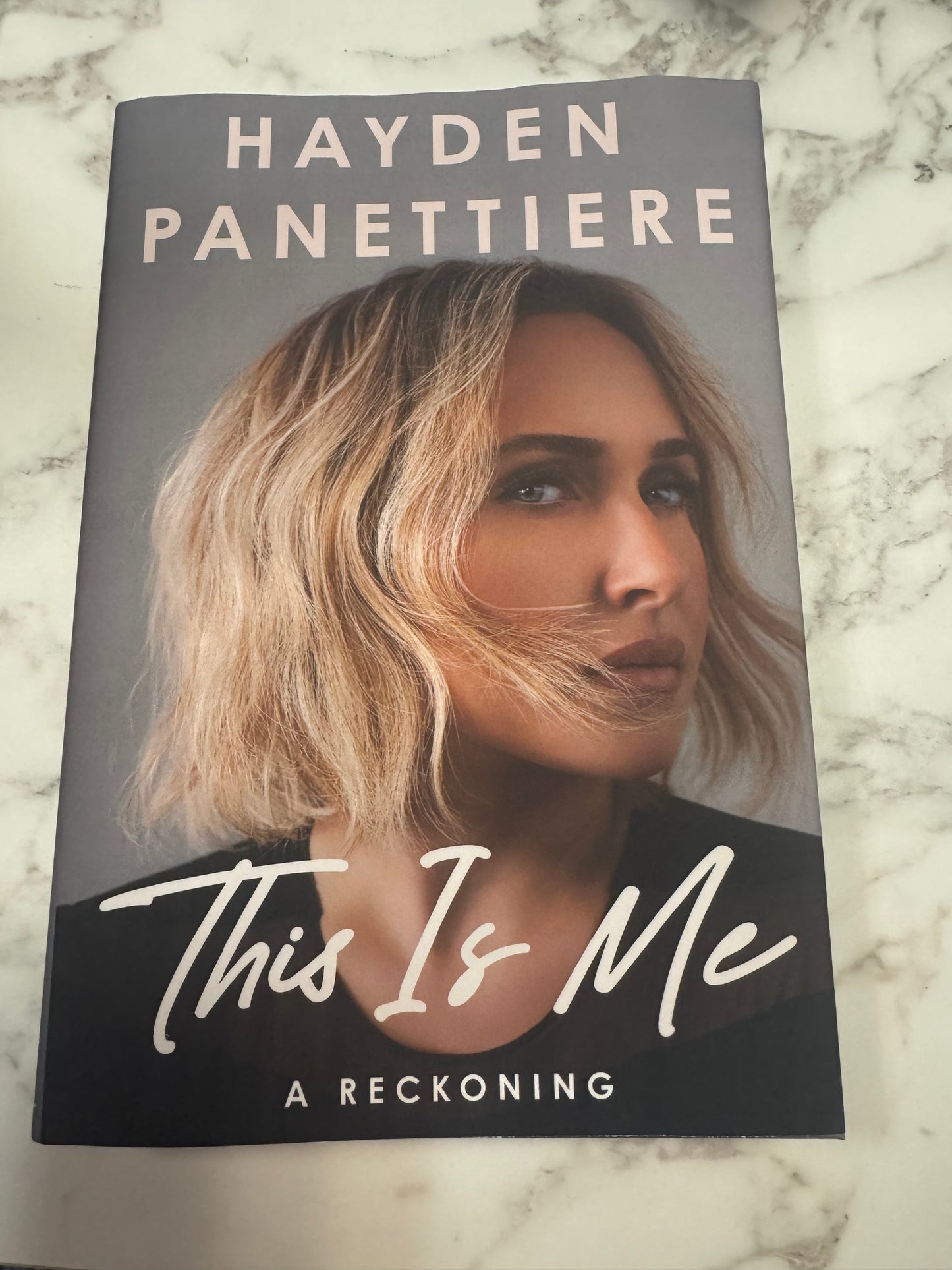 The front cover of the book 'This Is Me: A Reckoning' by Hayden Panettiere, featuring a portrait of the author on a gray background, placed on a white and gray marble surface.