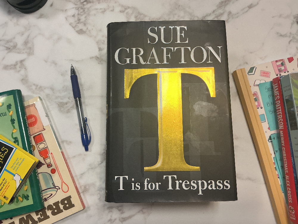 Book titled 'T is for Trespass' by Sue Grafton on a marble surface with other books and a pen.