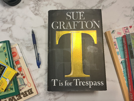 Book titled 'T is for Trespass' by Sue Grafton on a marble surface with other books and a pen.