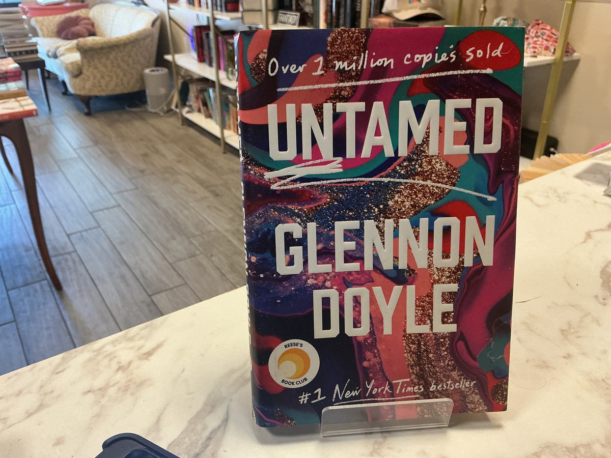 The book 'Untamed' by Glennon Doyle stands upright on a small clear display stand on a marble counter inside a bookstore. The cover features a vibrant, abstract multicolor design with pink, purple, blue, and gold glitter textures, and bold white text.