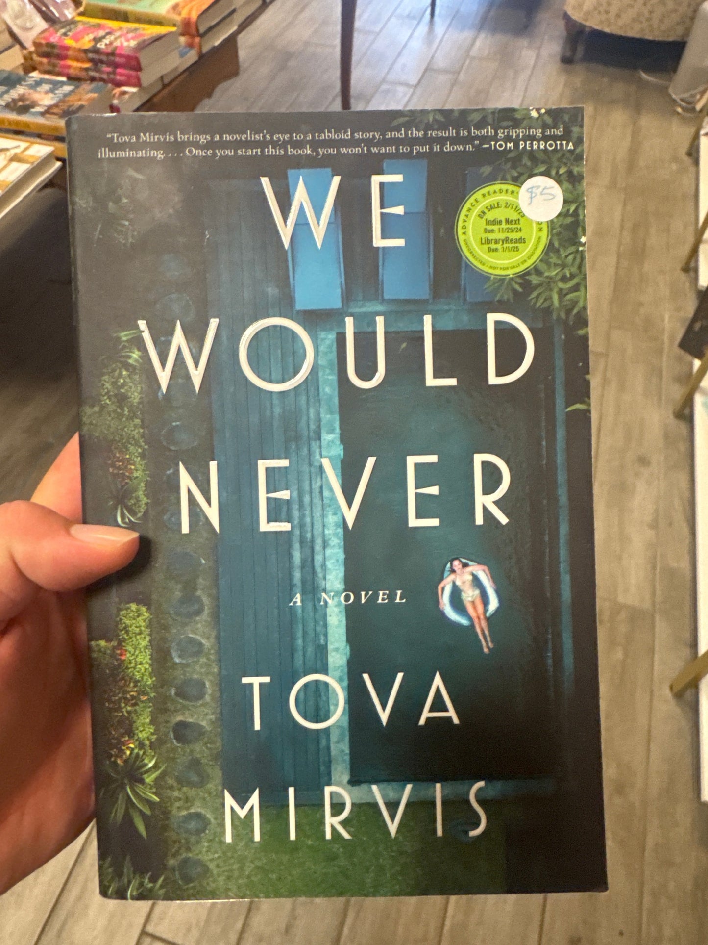 A hand holding a paperback copy of the novel 'We Would Never' by Tova Mirvis, photographed in a bookstore with wooden floors and bookshelves visible in the background.