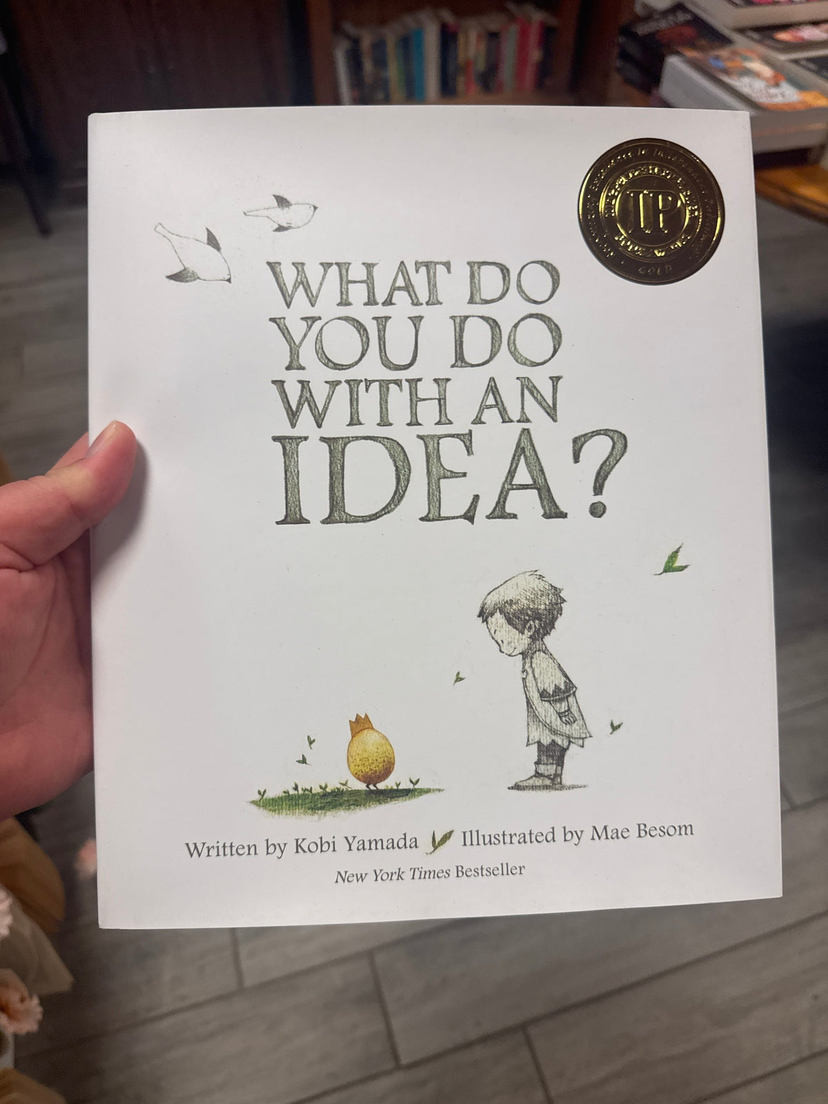 A hand holding a copy of the children's book 'What Do You Do With an Idea?' by Kobi Yamada, illustrated by Mae Besom. The white cover features gray title text, a small yellow bird, and a child illustration, with a gold New York Times Bestseller seal in the top right corner.