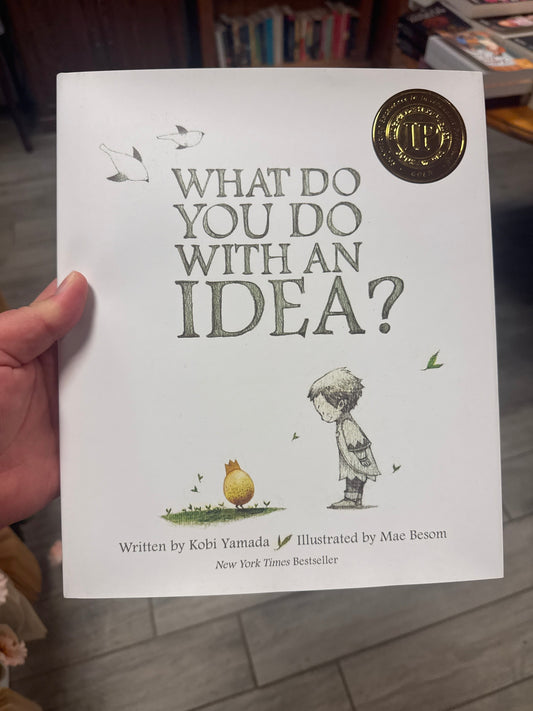 A hand holding a copy of the children's book 'What Do You Do With an Idea?' by Kobi Yamada, illustrated by Mae Besom. The white cover features gray title text, a small yellow bird, and a child illustration, with a gold New York Times Bestseller seal in the top right corner.