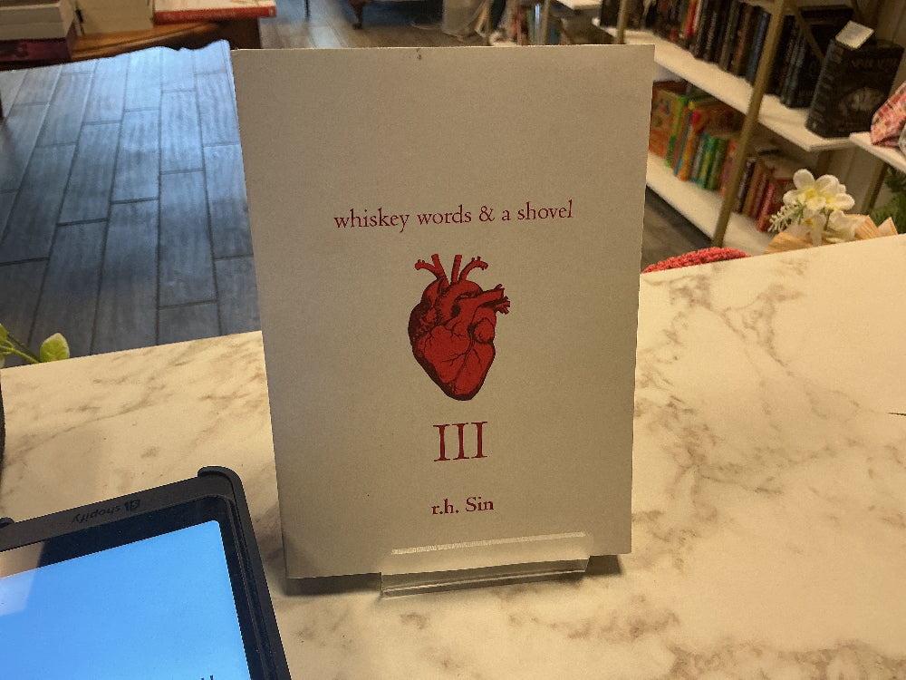 A book cover or booklet titled "whiskey words & a shovel" with a red anatomical heart illustration and the Roman numeral III, displayed on a marble counter in a bookstore or library setting.