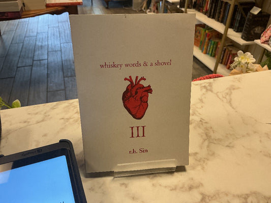 A book cover or booklet titled "whiskey words & a shovel" with a red anatomical heart illustration and the Roman numeral III, displayed on a marble counter in a bookstore or library setting.