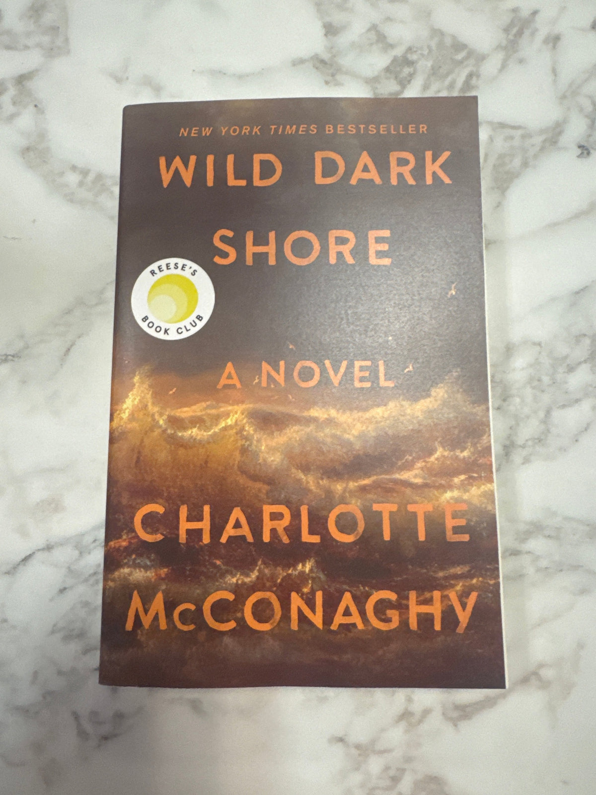 A paperback copy of the book 'Wild Dark Shore' by Charlotte McConaghy, featuring a dark brown and orange cover with a Reese's Book Club sticker, placed on a white marble surface.